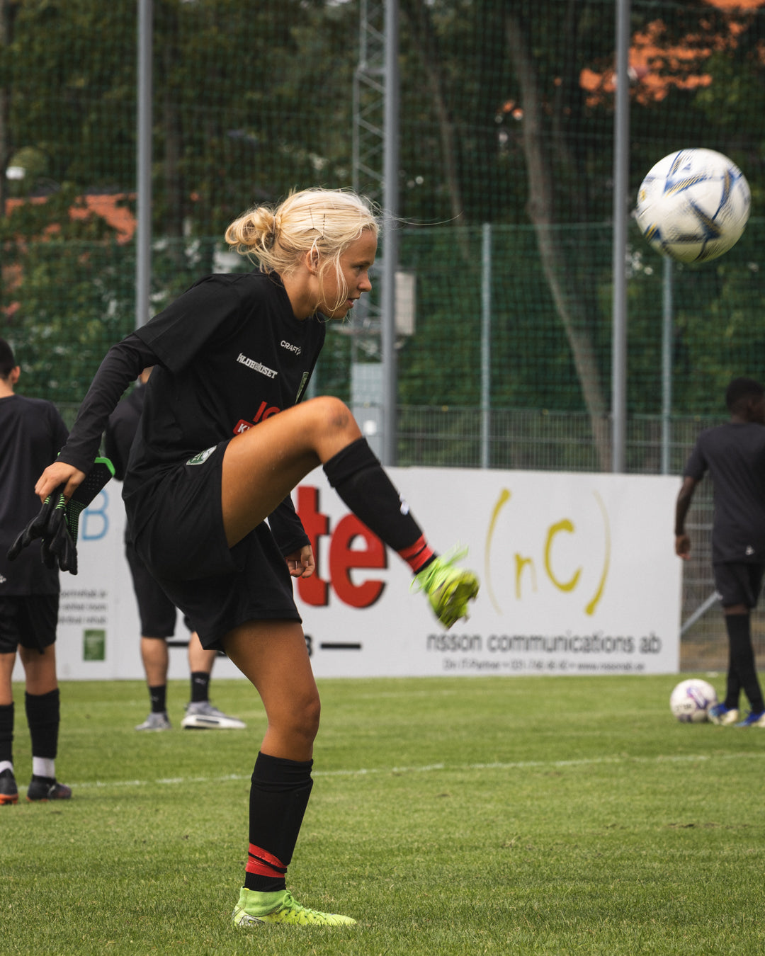 Woman in black sports outfit kicking a soccer ball on a field with advertisements in the background